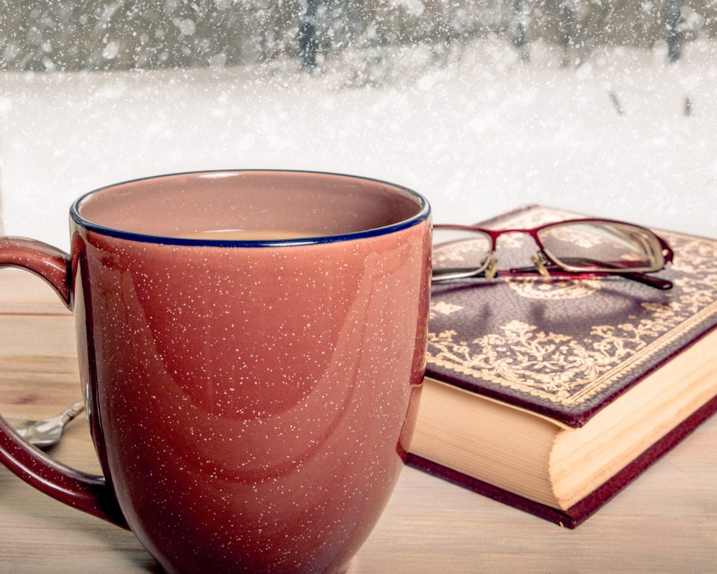 Cozy winter scene with coffee mug, book and snowfall symbolizing mindful holidays and stress relief.